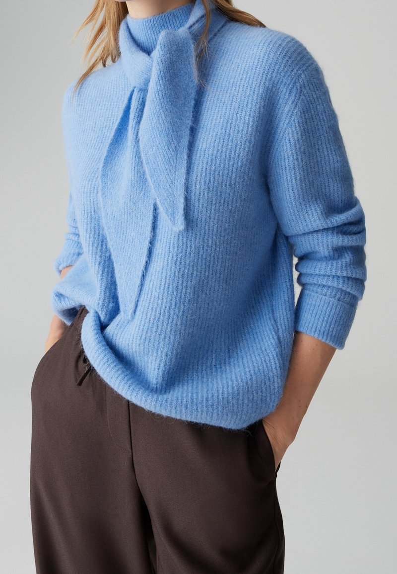 Personne portant un pull en maille bleu clair avec un détail d'écharpe nouée, associé à un pantalon ample marron foncé, mains dans les poches.