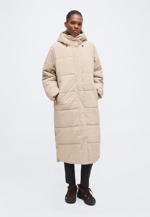 Femme portant un long manteau d'hiver matelassé beige avec capuche, pantalon noir et chaussures de randonnée noires avec des lacets rouges, debout les mains dans les poches.
