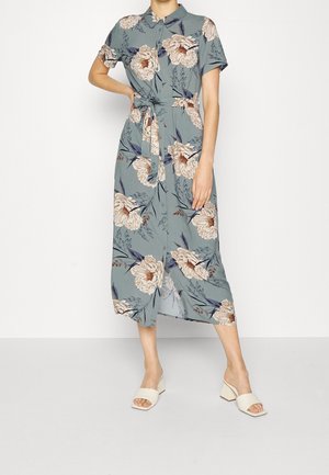 Kobieta w niebieskiej, kwiecistej sukience midi z krótkimi rękawami, wiązaną w talii i zapiętą na guziki z przodu, w połączeniu z białymi sandałami na obcasie.