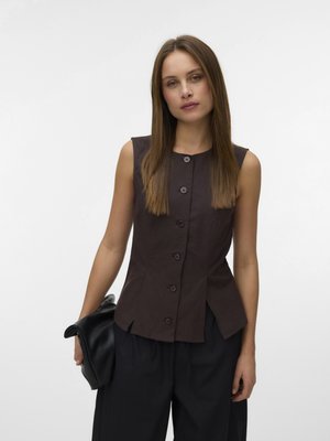 Nő egyenes barna hajjal, ujjatlan sötétbarna begombolt felsőt és bő fekete nadrágot visel, egy összecsukott fekete bőr táskát tartva.