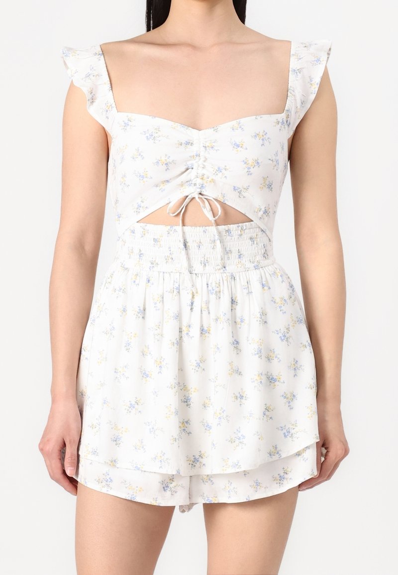 Robe florale blanche avec des manches courtes à volants, design découpé et taille smockée. Présente de petits motifs floraux bleus et jaunes.