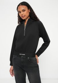 Čierna mikina na zips s kapucňou, elastický pás s logom "JOOP! JEANS", spárovaná s tmavými džínsami a hladkou textúrou.