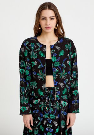 Kvinde iført sort cropped jakke og matchende nederdel med grøn og blå tropisk blomsterprint, stående mod en ensfarvet hvid baggrund.