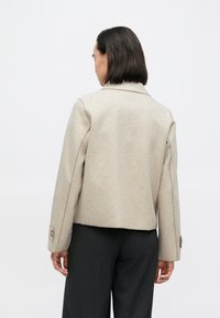 Beige jakke av ullblanding med firkantet fasong, hakket krage og knappede mansjetter, kombinert med mørke bukser, sett bakfra.