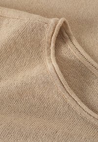 Tessuto a maglia beige con una texture liscia, caratterizzato da un semplice scollo rotondo e dettagli di cucitura visibili lungo i bordi.