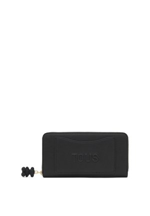 Cartera rectangular negra con cierre alrededor, con el logo TOUS en relieve y cremallera con colgante en forma de oso sobre fondo blanco.