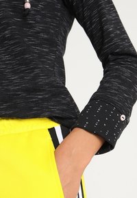 Person som bär en svart långärmad skjorta med mönstrade manschetter och ljusa gula byxor med ett svart och vitt randigt midjeband, hand i fickan.