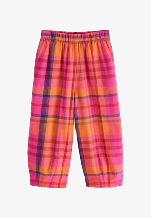 Niña joven con camisa blanca de gran tamaño, pantalones de cuadros rosa brillante y naranja, sosteniendo una chaqueta de mezclilla azul, de pie sobre un pie.