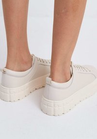 Beige lage sneakers met een glad oppervlak, een dikke witte zool met een gestructureerd patroon en een klein trekhaakje aan de achterkant.