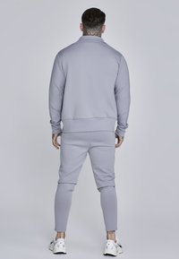 Completo sportivo grigio chiaro composto da una felpa con zip e colletto e pantaloni sportivi a gamba stretta. Il materiale sembra morbido e liscio. Le sneakers presentano dettagli neri.