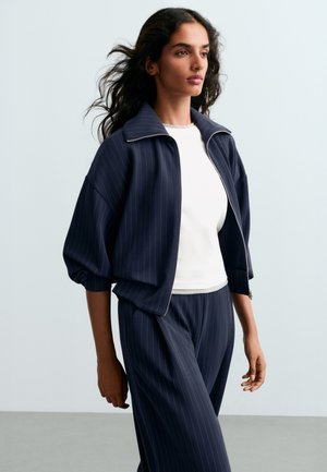 Femme aux longs cheveux foncés porte une veste zippée à rayures bleu marine et un pantalon assorti par-dessus un haut blanc, debout devant un fond pâle.