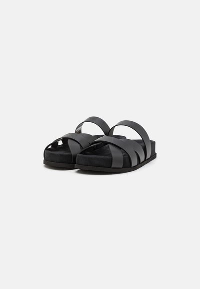 Sandales noires à glissière avec des sangles en synthétique lisse dans un design en croix, dotées d'une semelle noire douce et d'une esthétique minimaliste.