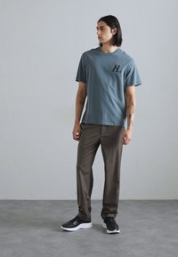 Gråblå t-shirt med kort ärm och svart 'HL'-logotyp, kombinerad med bruna byxor och svarta sneakers, stående på en grå golv mot en vägg.