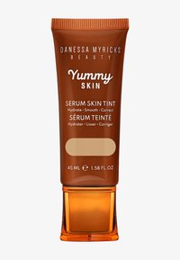 Brun tube med en klar orange bas, märkt "Yummy Skin Serum Skin Tint", 45 mL. Har text i vitt och ljusbeige. Slät textur.