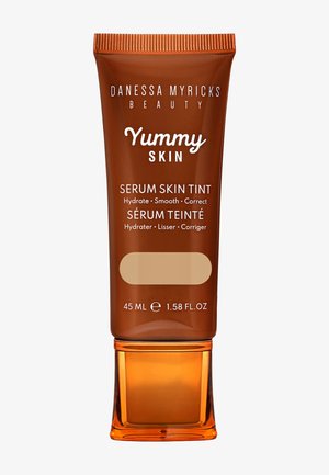 Bruine tube met een heldere oranje basis, gelabeld "Yummy Skin Serum Skin Tint," 45 mL. Bevat tekst in wit en lichtbeige. Gladde textuur.