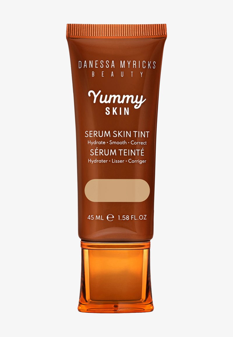 Brun tube med en klar orange bas, märkt "Yummy Skin Serum Skin Tint", 45 mL. Har text i vitt och ljusbeige. Slät textur.