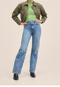 Olivgrüne cropped Jacke, gemustertes grünes Oberteil, hellblaue Schlagjeans, schwarze Knöchelstiefel; lässiges Outfit mit gemischten Texturen und Farben.