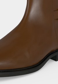Gros plan d'une botte cheville en cuir marron lisse avec semelle noire et petit talon empilé sur fond blanc.