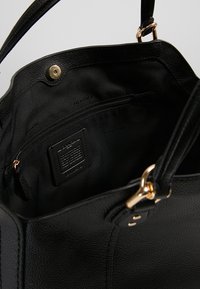 Sac à main en cuir noir avec des garnitures dorées, montrant la doublure intérieure en tissu, une poche zippée et un patch de marque en cuir.