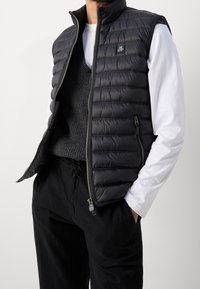 Hombre vistiendo un chaleco acolchado negro sobre un suéter de punto negro y una camisa blanca de manga larga, con pantalones negros, manos en los bolsillos, sobre un fondo liso.