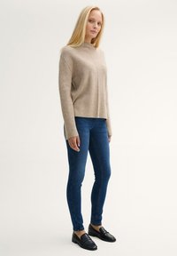 Suéter beige con un cuello alto y hombros caídos, combinado con jeans skinny azul oscuro y mocasines negros. Textura suave, diseño minimalista.