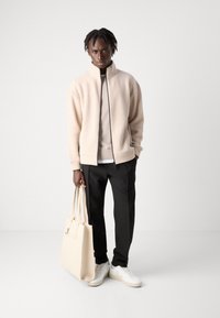 Beige fleece jacket with high collar, black pants, white shoes, and beige tote bag. Smooth texture and minimalistic design.