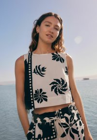 Zwart-witte cropped top met bloem- en bladmotieven, voorzien van een effen zwarte sectie en decoratieve witte knopen langs de zijkant.