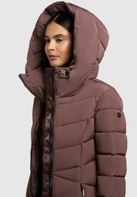 Hnědá péřová bunda s velkou kapucí, obsahující přední zip, prošívané detaily a božní štítek s logem. Hladká, prošívaná textura.