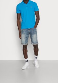Polo bleu avec col et logo Lacoste, associé à un short en denim clair, des baskets blanches et des chaussettes mi-mollet rayées.