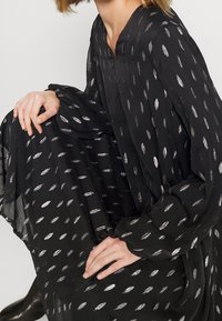 Robe noire en tissu léger avec un décolleté en V, ornée de motifs en points argentés et de manches longues. Texture douce avec un design fluide.