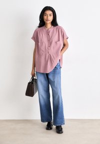 Ροζ ριγέ κοντομάνικο πουκάμισο με στρογγυλή λαιμόκοψη και δύο μπροστινές τσέπες, συνδυασμένο με φαρδιά μπλε τ�ζιν και μια υφασμάτινη καφέ τσάντα.