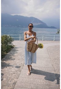 Abito senza maniche di colore azzurro chiaro; borsa in paglia intrecciata con manici marroni; infradito; sentiero all'aperto vicino a un lago, con montagne sullo sfondo.