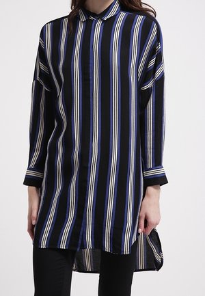 Vrouw die een tuniek met lange mouwen en kraag draagt, met verticale zwarte, blauwe en witte strepen, gecombineerd met een zwarte broek.