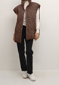 Brązowa pikowana kamizelka bez rękawów z wysokim kołnierzem, z zatrzaskami, noszona na białej bluzce z długim rękawem i połączona z czarnymi spodniami.