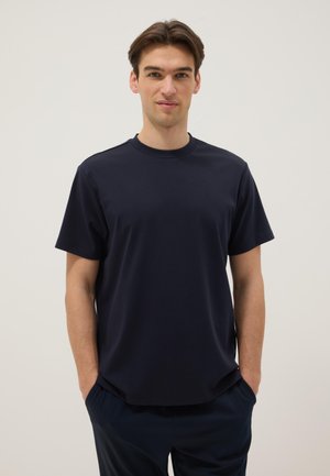 Joven con cabello castaño que lleva una camiseta de manga corta azul marino oscura lisa y pantalones oscuros, de pie con las manos en los bolsillos contra un fondo blanco.