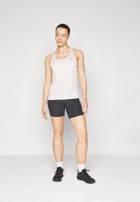 Lyserød ærmeløs tanktop med et lille logo, parret med sorte tætsiddende shorts. Modellen bærer sorte sportsko og hvide ankelsokker.