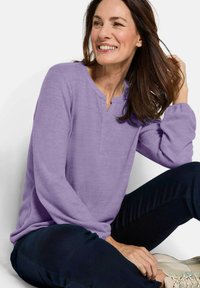 Lila Pullover mit langen Ärmeln, einem V-Ausschnitt und hochgekrempelten Ärmeln, kombiniert mit dunklen Hosen. Weiche Textur und einfaches Design.