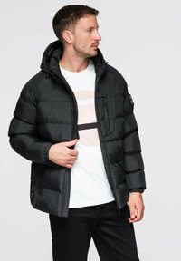 Schwarze gesteppte Jacke mit Kapuze, mit wattierter Textur, Reißverschlusstasche auf der Brust, elastischen Bündchen und einem schlanken Design. Kombiniert mit einem weißen Hemd.