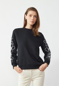 Svart hettegenser med lange ermer, prydet med hvite blomsterbroderier langs ermene. Casual passform med rund hals og ribbestrikkede mansjetter.