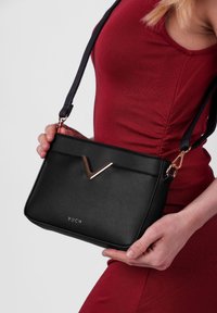 Černá kožená kabelka s vertikálním zipem, zlatým V-tvarovaným akcentem a nastavitelním popruhem. Název značky "VUCH" vyražený na přední straně.