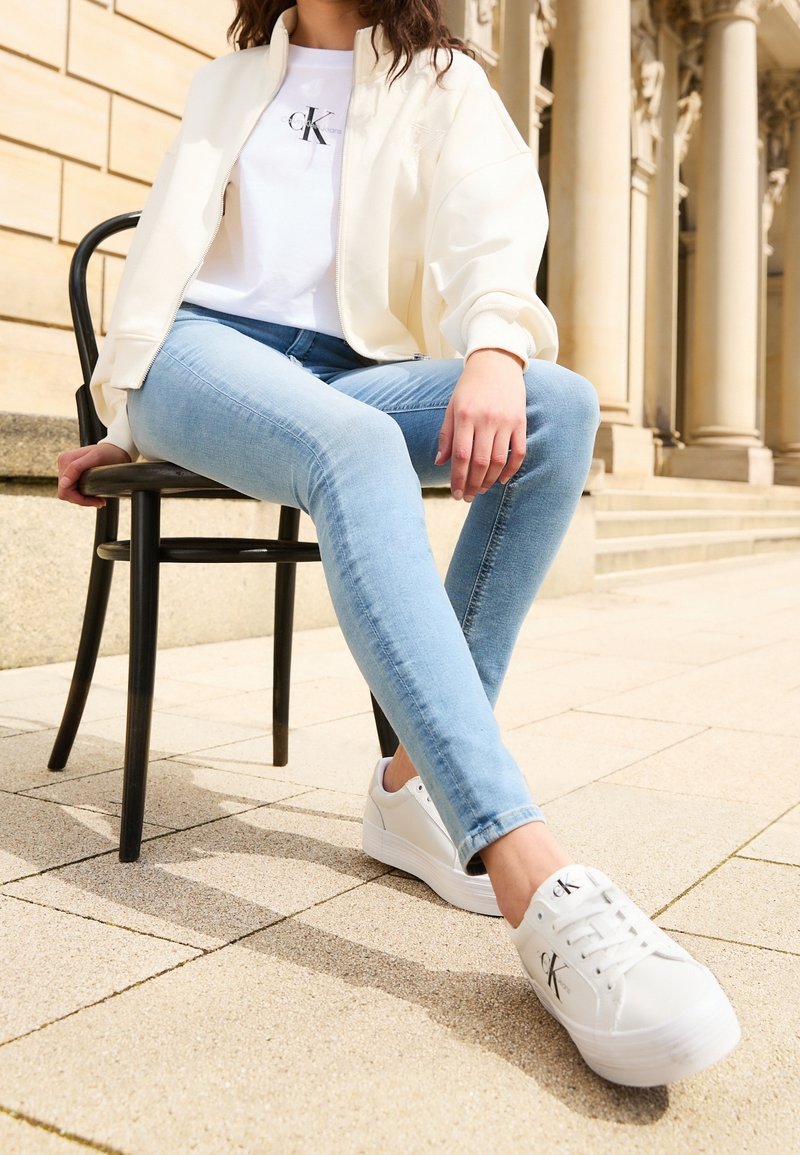 Vita sneakers med tjock sula och svart logotyp, ljusblå skinny jeans och en off-white bomberjacka över en vit T-shirt.