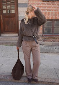 Jachetă maro din blană sintetică cu centură, pantaloni carouri bej, și geantă tote din piele întoarsă maro închis, pe fundalul unui zid de cărămidă și a unei uși din lemn.