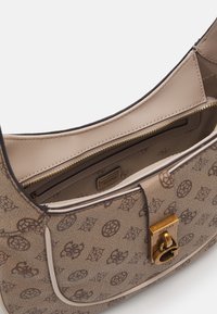 Béžová kabelka Guess s hnědým logem, zlatým zapínáním a krémovým vnitřkem, který obsahuje zipovou přihrádku s označením Guess.