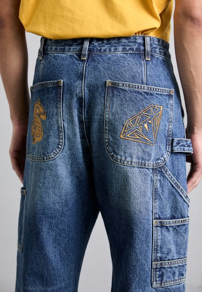 Pantalons cargo en denim, bleu clair avec des coutures visibles. Comprend des poches arrière avec une broderie dorée d'un dragon et d'une forme de diamant.
