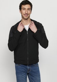 Jachetă neagră cu fermoar, cu accente texturate pe umeri. Prezintă un design ajustat și manșete răsucite. Purtați-o peste un tricou alb și blugi albaștri.