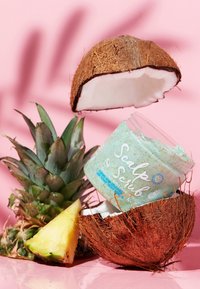 Scalp scrub in een doorzichtig potje met mintgroene scrub, omringd door een gehalveerde kokosnoot en een ananasschijf, tegen een roze achtergrond.
