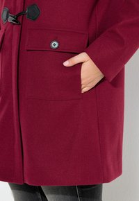 Cappotto in lana borgogna con grandi tasche frontali, chiusure con bottoni neri e una texture liscia. Mano posizionata nella tasca per scala.