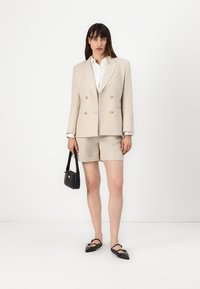 Béžový lněný dvouřadý blazer se zlatými knoflíky v kombinaci s odpovídajícími šortkami. Černá kabelka a špičaté ploché boty dotvářejí vzhled.