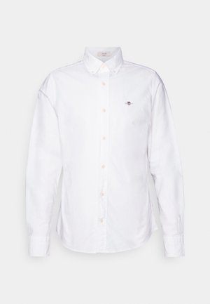 White button-up shirt made of cotton. Features a pointed collar, long sleeves, and light tan buttons. Small embroidered logo on the chest.