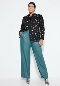 Chemise noire à boutons, ornée de motifs circulaires colorés, associée à un pantalon large en teal pailleté et des escarpins argentés à bout pointu.